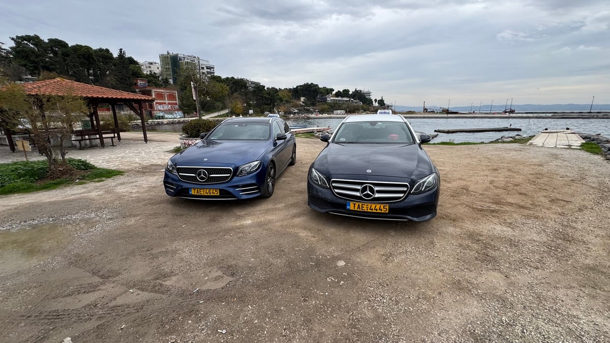 Two Mercedes-Benz sedans parked on a sandy beach with waterfront and boats visible in the background